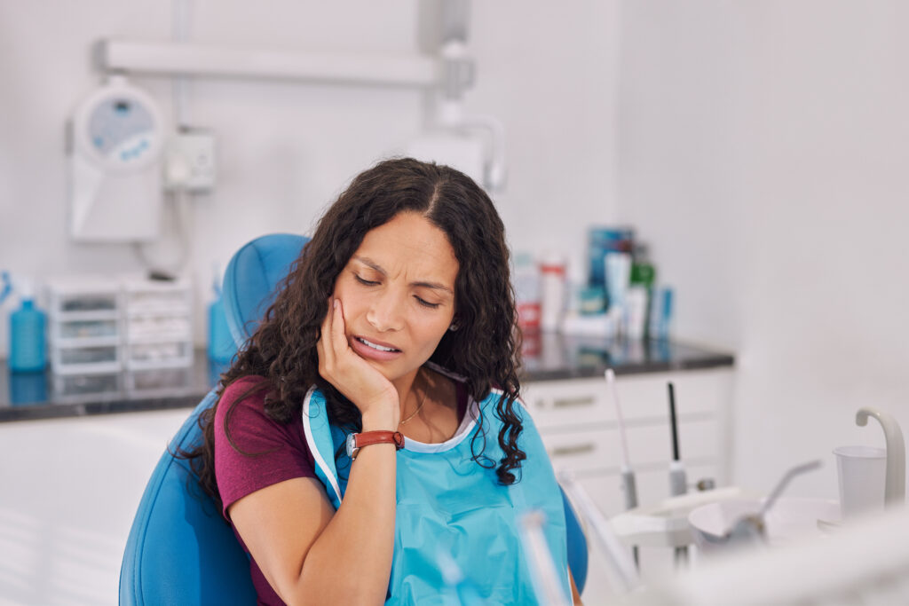 Young mixed race woman sitting in dental chair holding jaw in discomfort. Patient showing signs of tooth pain during consultation, oral infection or caria. Woman wearing dental bib in clinic with hands on face pressing her cheek, showing discomfort from a toothache.