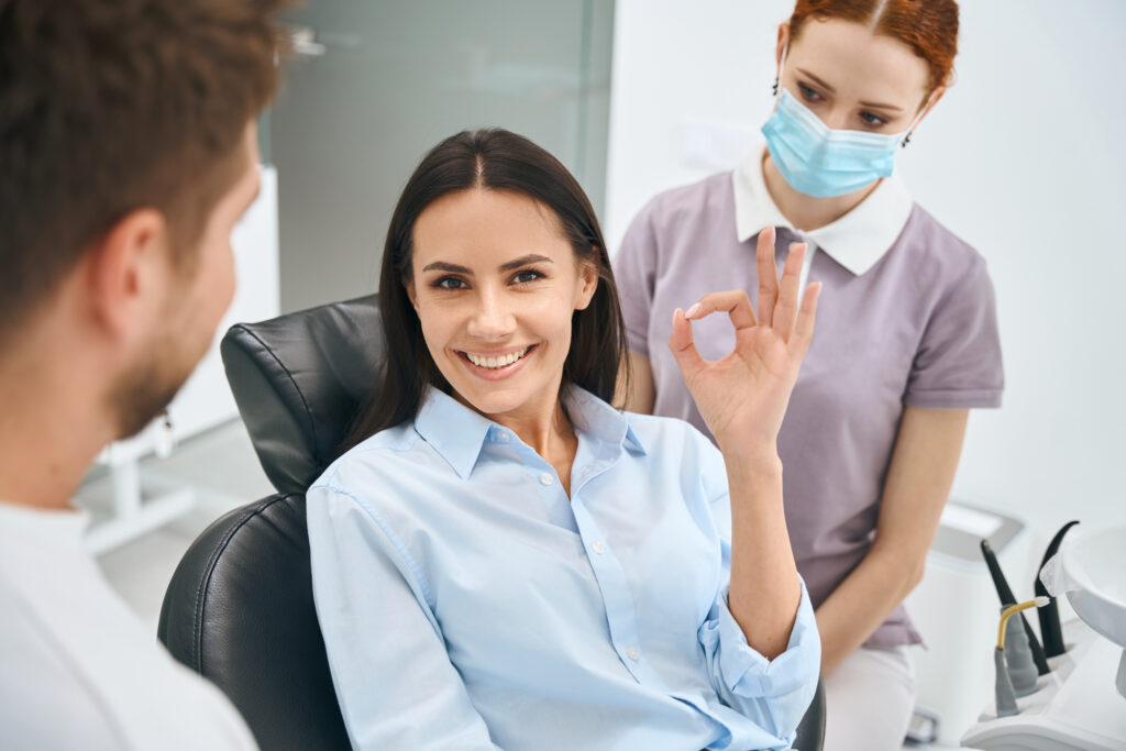 Smiling beautiful woman showing ok gesture sign sitting in orthodontic chair after painless and quality dental treatment, qualified dentist and nurse standing nearby
